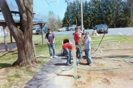 Four scouts are working on getting posts in the ground.  The one on the left is standing with his hands in his pockets.  The one on the center left is bending down filling a bucket with water.  The one on the center right is digging with the post-hole digger.  The one to the right is staring at the camera, but holding a water bottle up to his face so you can't see him.